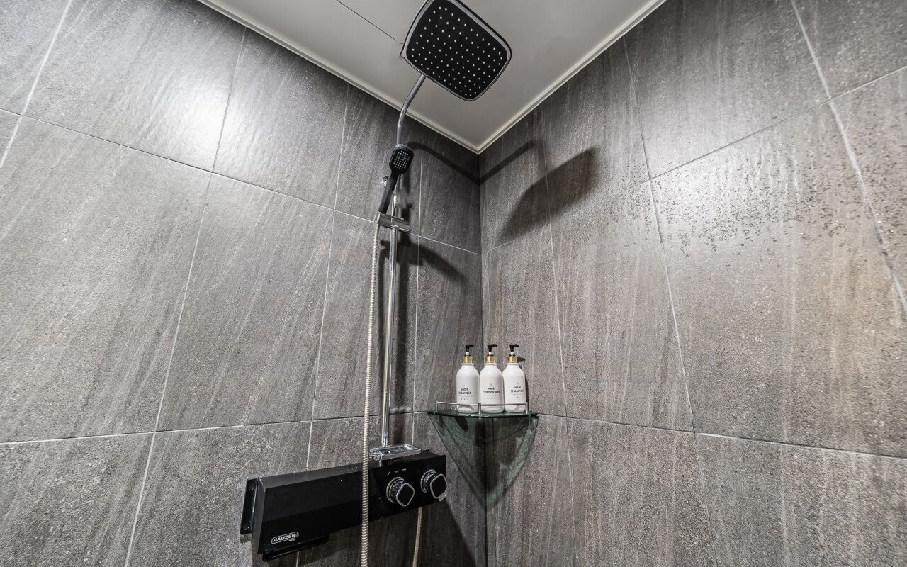 A modern shower area with a rain showerhead, a handheld showerhead, and a control panel. There are three bottles on a shelf above the shower.