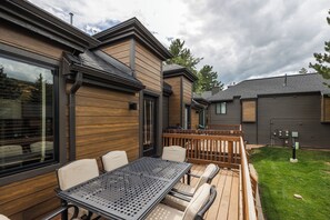 A patio area with a table and chairs, a wooden deck, and a view of a house with large windows and a dark roof. - Canyons Retreat: Alpine Charm, Shared Hot Tub/Pool, Sleeps 10, Cozy Fireplace (Park City)
