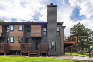 A modern multi-story residential building with a prominent chimney, multiple windows, and a wooden deck. - Canyons Retreat: Alpine Charm, Shared Hot Tub/Pool, Sleeps 10, Cozy Fireplace (Park City)
