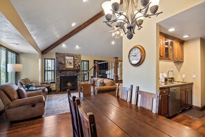 A spacious living area with a fireplace, dining table, and seating. A kitchen with wooden cabinets and a clock is visible. - Canyons Retreat: Alpine Charm, Shared Hot Tub/Pool, Sleeps 10, Cozy Fireplace (Park City)