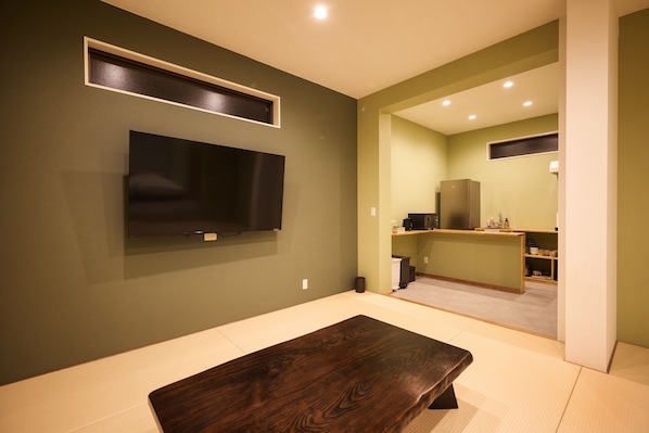 A modern living room with a flat-screen TV mounted on the wall, a wooden coffee table, and a kitchen area visible through an open doorway. - Adan Resort  NAGOMI (Motobu)