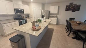 Une cuisine moderne avec des meubles blancs, des plans de travail en granit, un îlot central et un coin repas comprenant une table et des chaises en bois.