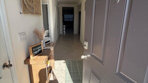 Un couloir avec une porte sombre, un petit banc en bois sur lequel est posée une pancarte, une décoration murale en fibres tressées et un sol carrelé.