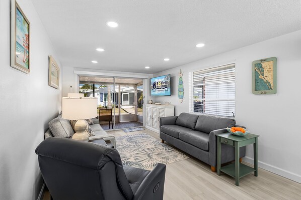 A modern living room with a grey sofa, a black armchair, a small green side table with a bowl of fruit, and a TV mounted on the wall. - Sun, Sand, and Relaxation! - Aloha Kai #43 - Roelens (Siesta Key)