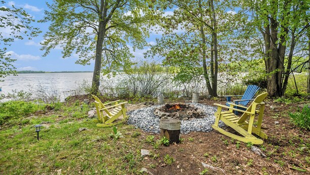 Un espacio al aire libre junto al lago con dos sillas Adirondack, un hogar para fuego y un banco de madera.