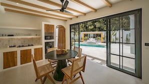 A modern kitchen with wooden cabinets, a dining table with chairs, and a large window overlooking a pool. - Palmera Aalto Bingin (Pecatu)