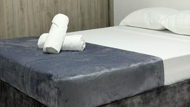 A bed with a grey mattress and white linens, a wooden wardrobe, and a white wall.