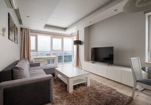 A modern living room with a grey sofa, a white coffee table, a TV on a white cabinet, and a view of the city through large windows. - Platinum Tower Skyline Apartment (Warszawa)