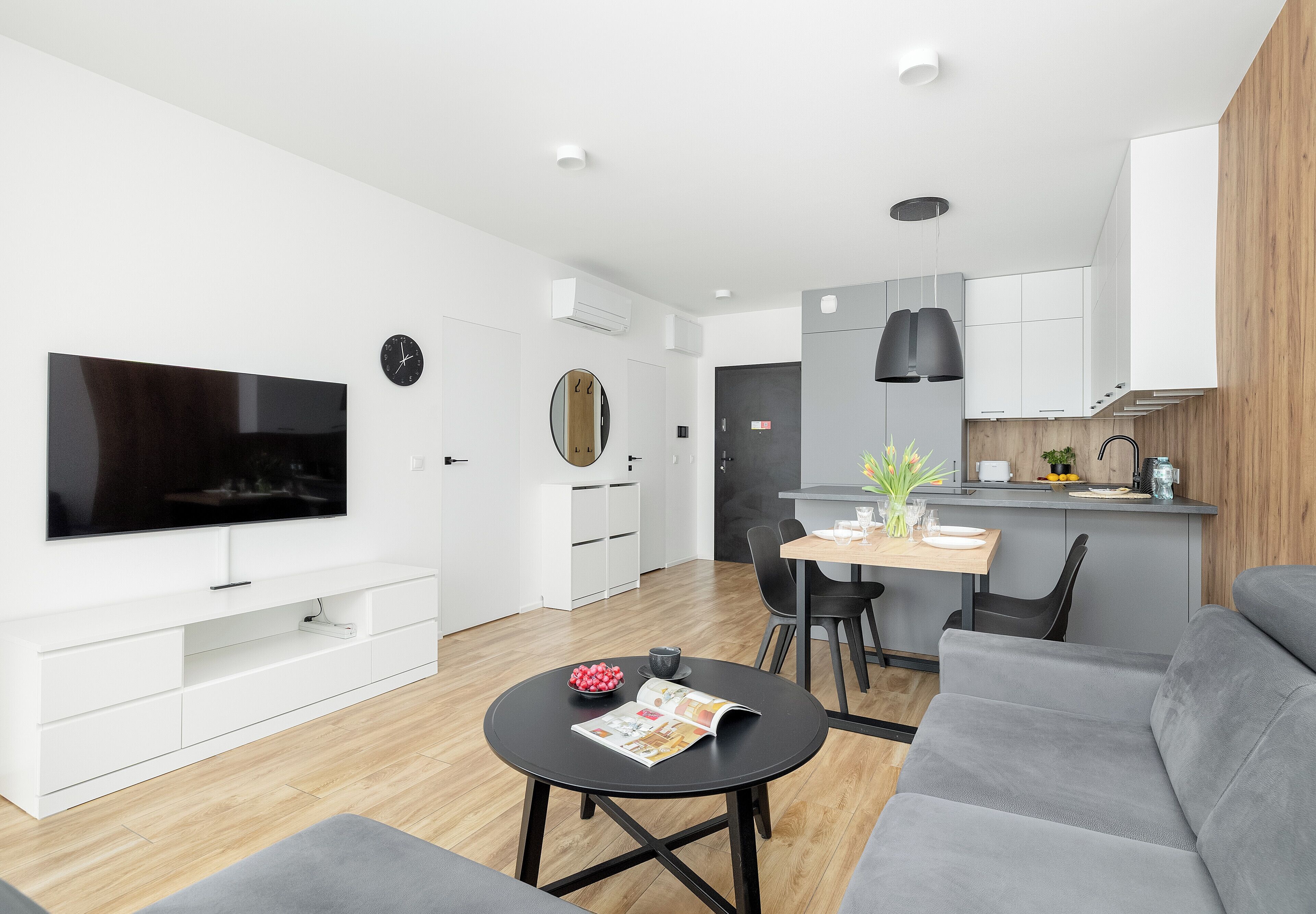 Un salon moderne avec un téléviseur à écran plat, une table basse ronde et un canapé gris. Il y a également un coin cuisine avec une table à manger et des chaises.