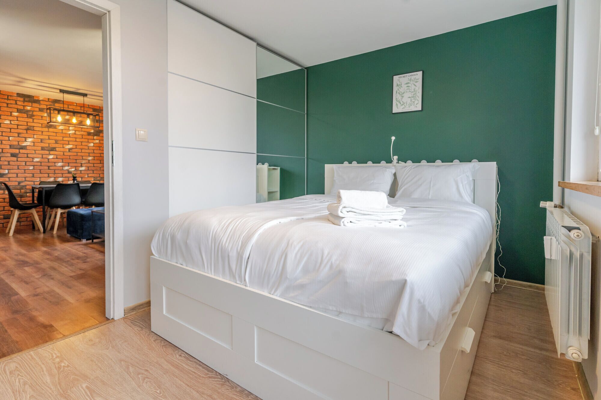 A neatly made bed with white linens, a green wall, and a brick wall in the background.