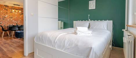 A neatly made bed with white linens, a green wall, and a brick wall in the background.