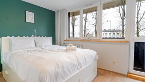 Una cama bien hecha en una habitación con paredes verde azulado, una ventana que da a vistas de árboles y edificios, y un pequeño cuadro en la pared.