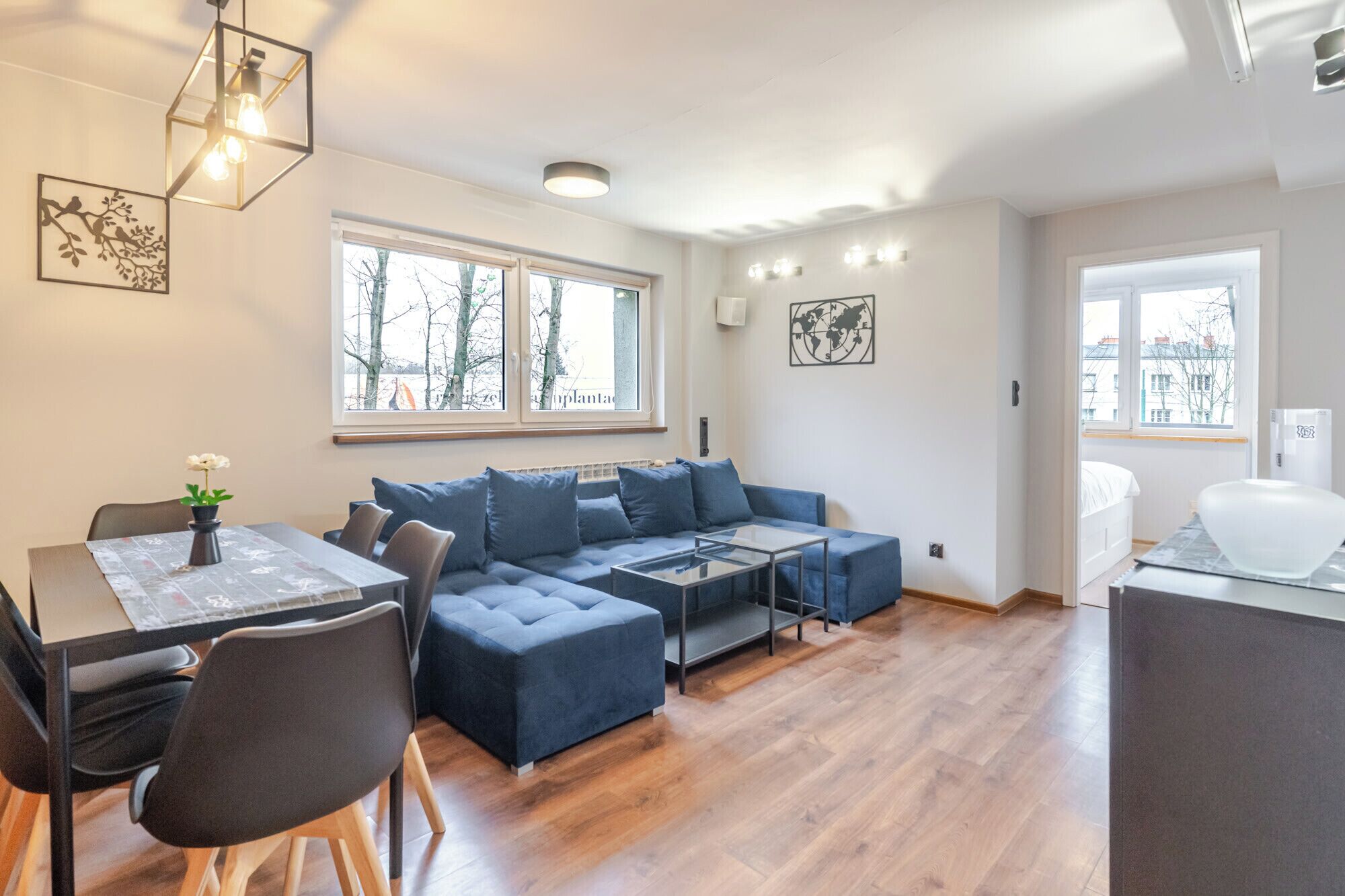 A modern living room with a sofa set, a dining table, and a coffee table. There are large windows providing natural light and views of the outside.