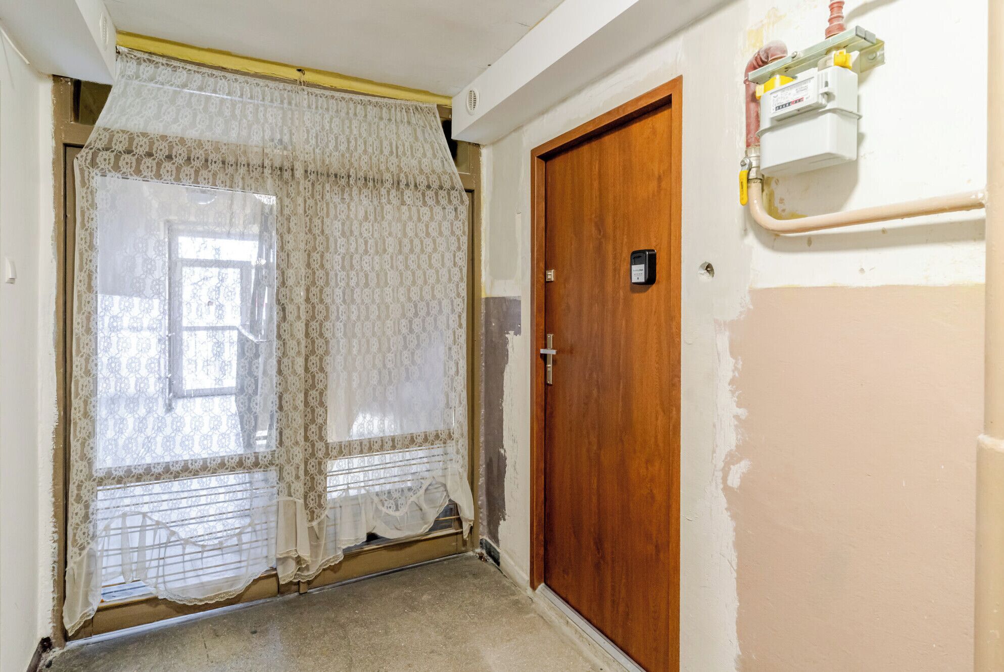 Un couloir étroit avec une porte en bois, une fenêtre ornée de rideaux en dentelle et un compteur d’électricité fixé au mur.