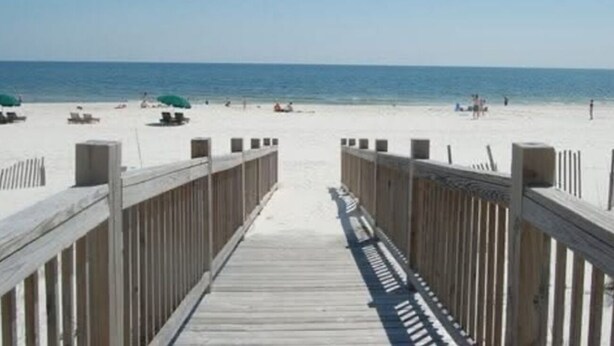 A wooden boardwalk leading to a beach with umbrellas and people in the distance. - Property is a great spot to relax in the sun with beach front and ocean view. (Gulf Shores)