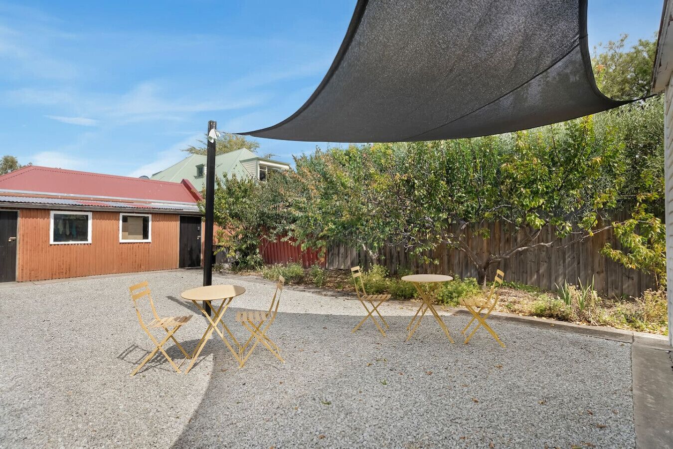 A gravel patio with a round table and four chairs, a black shade sail, a wooden fence, and a building with a red roof in the background.