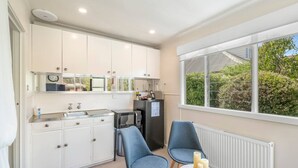 A compact kitchen with white cabinets, a microwave, and a refrigerator. There are two blue chairs and a small round table with candles. A window provides a view of greenery outside. - Private North Hobart Cottage (North Hobart)