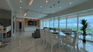 Un salon moderne avec une table à manger en verre, des chaises blanches et un grand téléviseur fixé au mur.