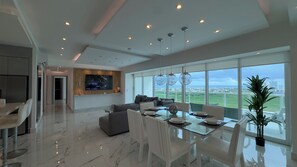 Ein modernes Wohnzimmer mit großem Fernseher, einem Essbereich mit Glastisch und einem Balkon mit Blick.