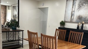 Un comedor con una mesa y sillas de madera, una estantería metálica negra con cristalería, y un cuadro abstracto de gran tamaño en la pared.