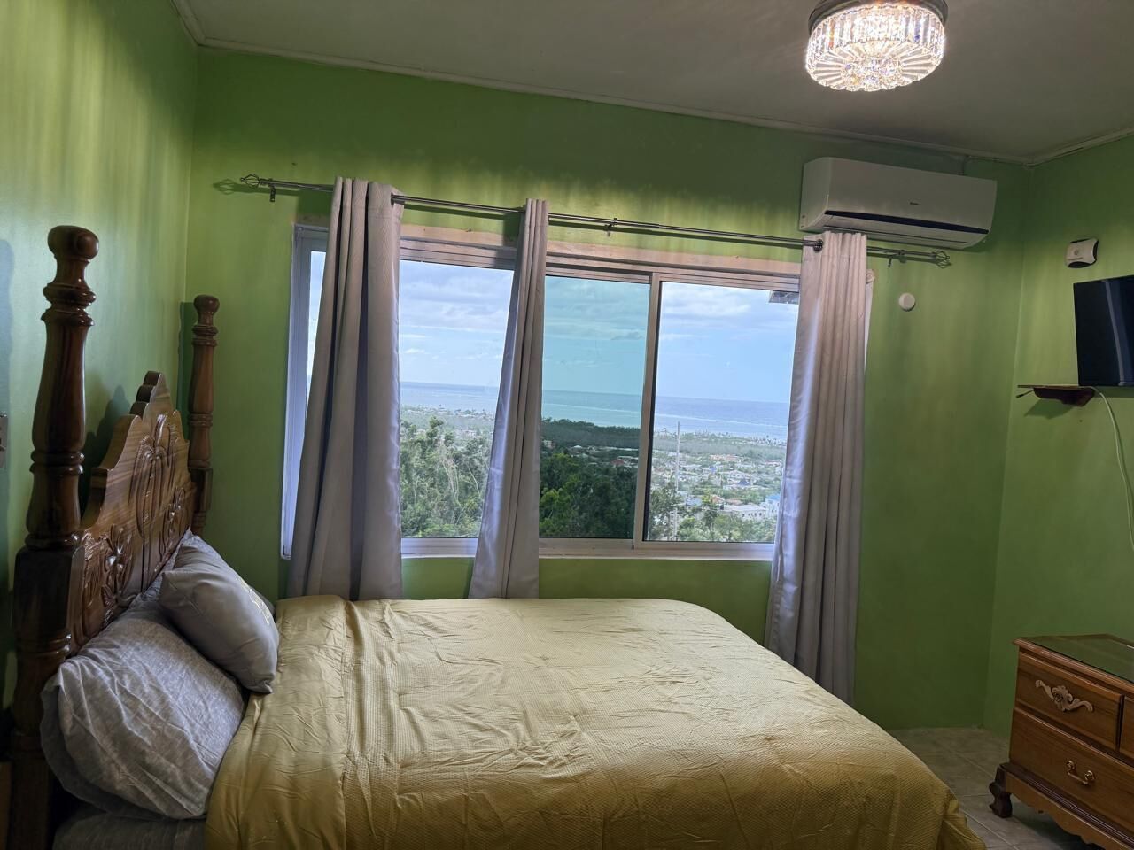 A bedroom with a bed, a wooden headboard, a dresser, an air conditioning unit, and a window with curtains.