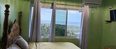 A bedroom with a bed, a wooden headboard, a dresser, an air conditioning unit, and a window with curtains.