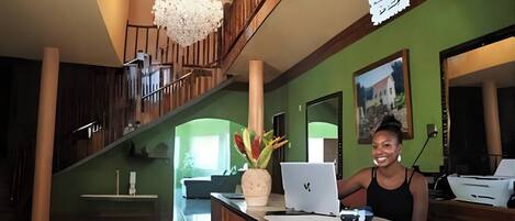 A reception desk with a laptop, a vase with flowers, and a staircase in the background.