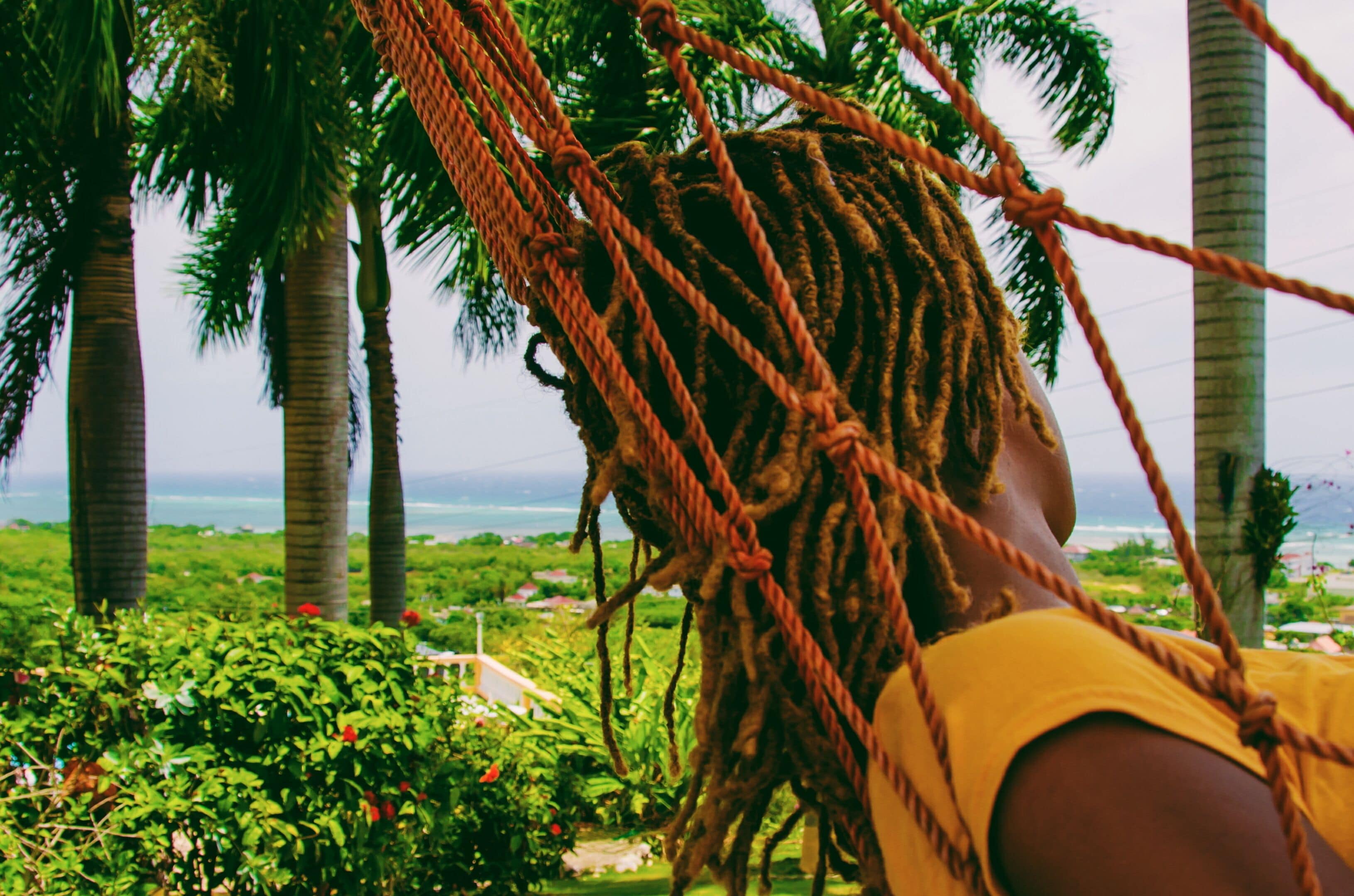 Una hamaca con cuerdas, una persona con rastas y vistas a palmeras y al océano.