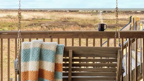 Un banc en bois, sur lequel est posée une couverture et une tasse, se trouve sur un porche donnant sur un paysage verdoyant.