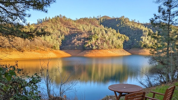 Blick auf einen See mit einem Holztisch und Stühlen, umgeben von Bäumen und Hügeln.