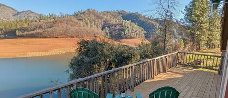 A wooden deck with two green chairs and a table, overlooking a lake and forested hills.