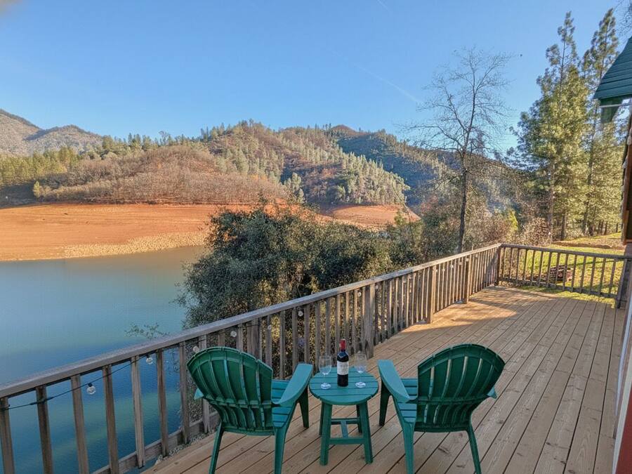Terraza de madera con dos sillas verdes y una mesa, con vistas a un lago y colinas boscosas.