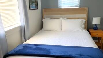 A bed with a wooden headboard, white bedding, and a blue blanket. There is a nightstand with a lamp and a picture on the wall.