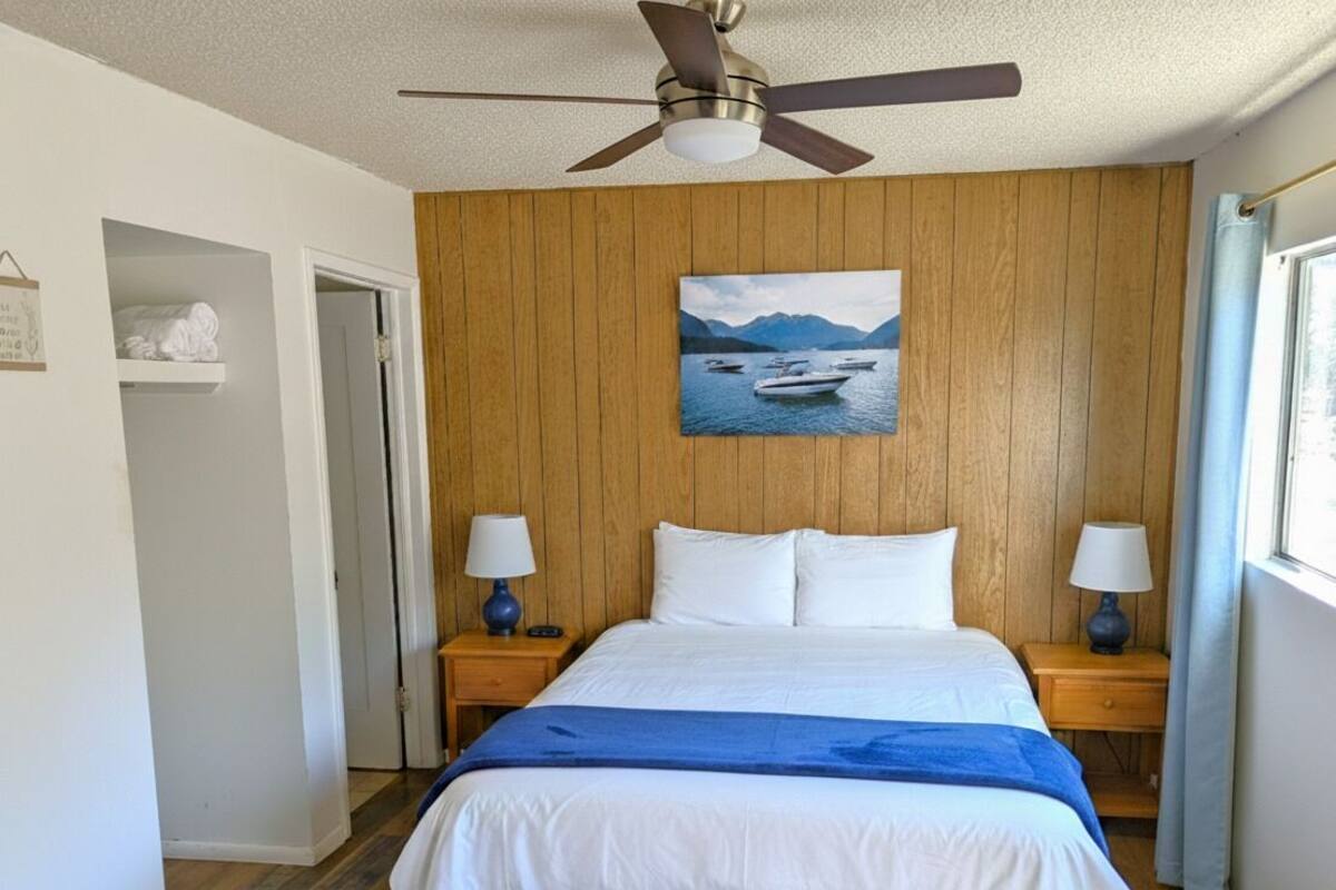 Une chambre à coucher avec un lit, des murs en bois, un ventilateur de plafond, un tableau représentant un lac et une fenêtre avec des rideaux.