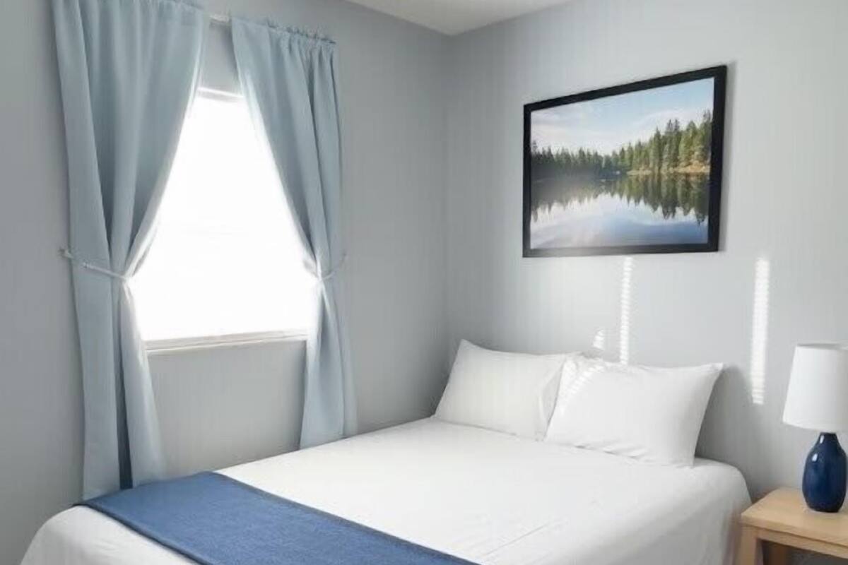 Un lit bien fait, agrémenté d’une touche de bleu, une table de chevet en bois avec une lampe, une fenêtre avec des rideaux et un tableau encadré au mur.