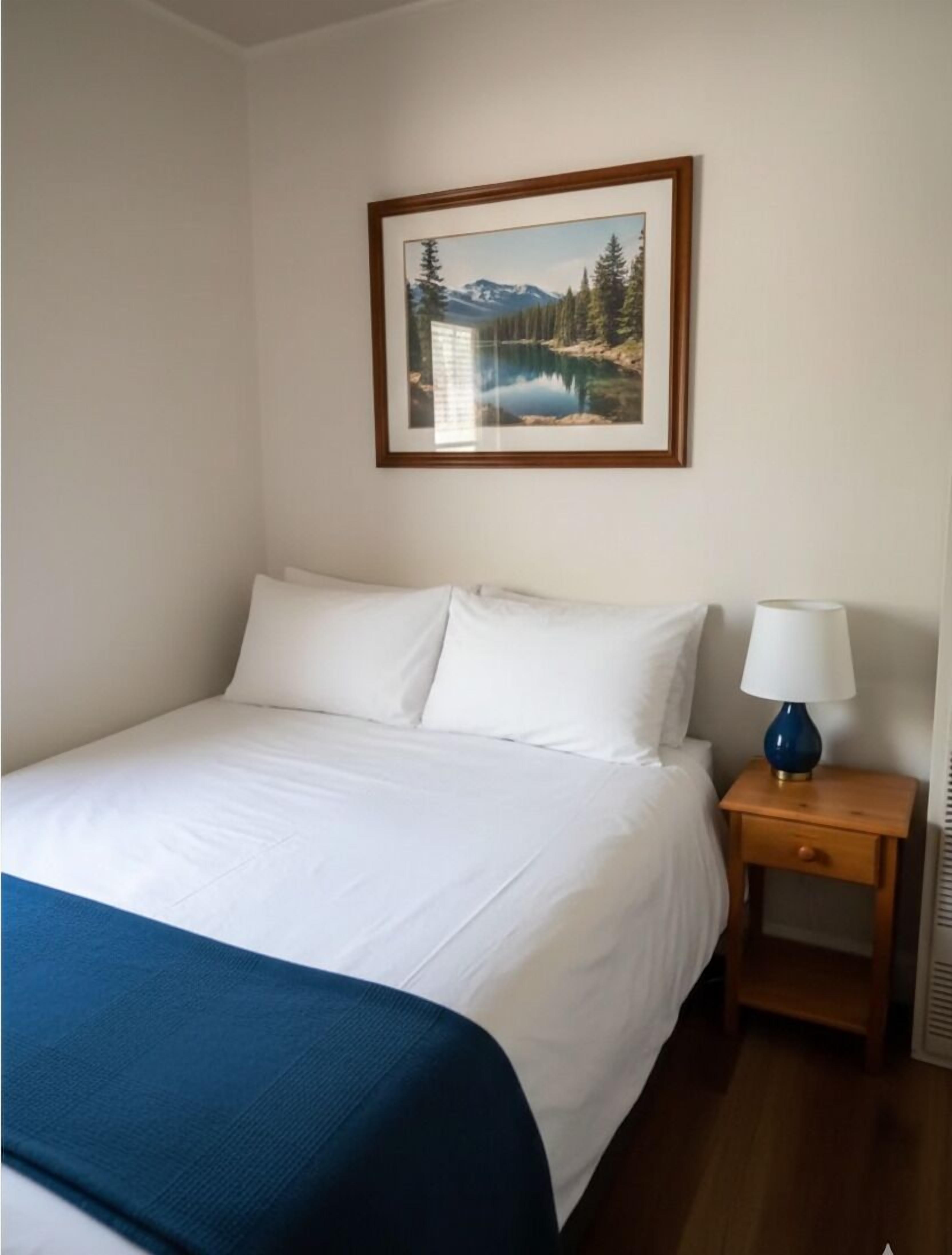 A neatly made bed with white linens and a blue throw blanket, a wooden nightstand with a lamp, and a framed picture of a mountain landscape on the wall.