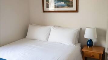 A neatly made bed with white linens and a blue throw blanket, a wooden nightstand with a lamp, and a framed picture of a mountain landscape on the wall.