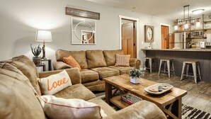 A cozy living room with a brown leather couch, a wooden coffee table, and a kitchen area with stools and a bar.