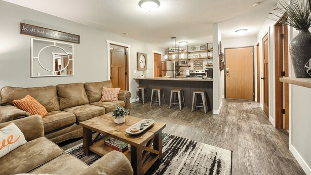 A modern living room with a brown leather sofa, wooden coffee table, and a kitchen area with stools and a bar counter.