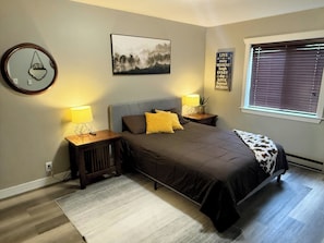 A bedroom with a bed, two bedside tables with lamps, a wall clock, a framed picture, and a window with blinds. - Black Hills Cozy Condo #2 (Lead)