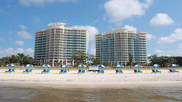 A beachfront hotel with two high-rise buildings, a sandy beach with blue lounge chairs, and a clear blue sky. - Amazing Gulf Views! Legacy Towers 10th Floor! (Gulfport)