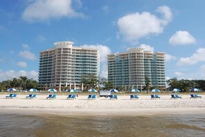 A beachfront hotel with two high-rise buildings, a sandy beach with blue lounge chairs, and a clear blue sky. - Amazing Gulf Views! Legacy Towers 10th Floor! (Gulfport)