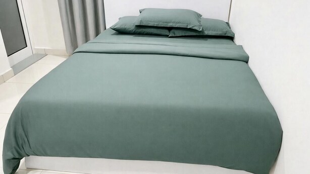 A bed with a green bedspread and pillows, a white headboard, and a grey curtain.