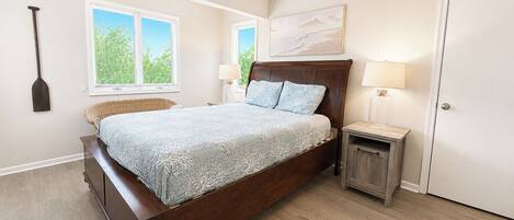 A bedroom with a wooden bed, two bedside tables, a window with blinds, a wall-mounted artwork, and a door.