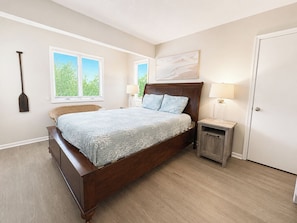 Ein Schlafzimmer mit einem hölzernen Bett, zwei Nachttischen, einem Fenster mit Jalousien, einem an der Wand befestigten Kunstwerk und einer Tür.