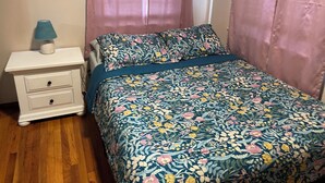 A bedroom with a floral bedspread, a white nightstand with drawers, a lamp, and pink curtains.