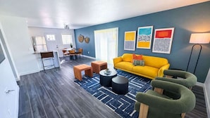 Una sala de estar moderna con un sofá amarillo, sillones verdes, una mesa de centro y un comedor con mesa y sillas.