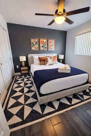 Un dormitorio con una cama, un ventilador de techo, un tapete geométrico y obras de arte en la pared.