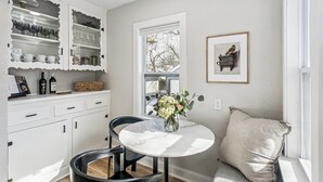 Una cucina con mobili bianchi, un tavolo di marmo rotondo, due sedie nere, un vaso con fiori, una finestra con vista e un quadro appeso al muro.