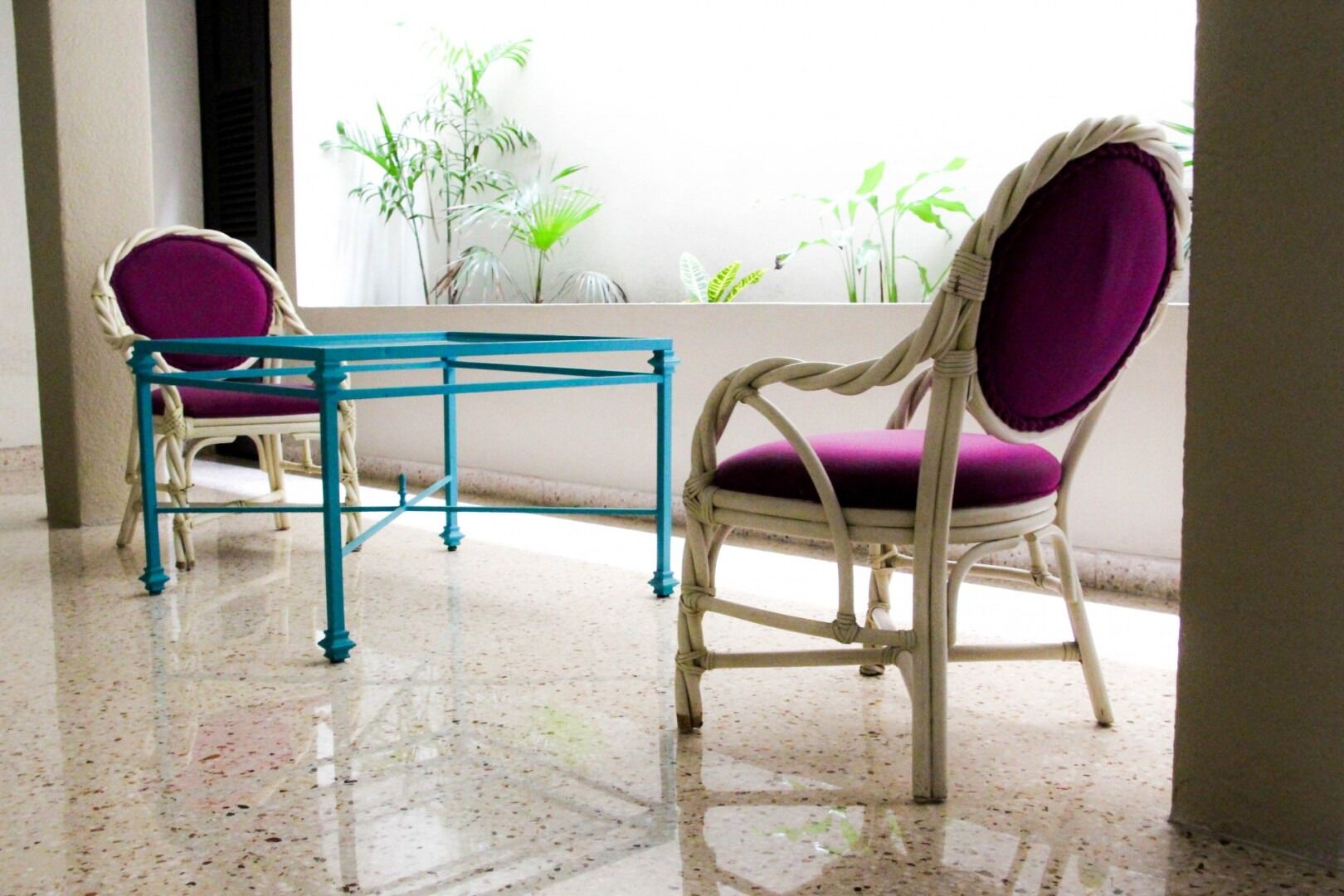 Duas cadeiras com almofadas roxas e uma mesa azul num quarto com chão de azulejos e uma janela com plantas.